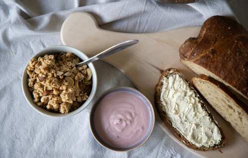 Brot auf Holzbrett neben Schüssel mit Joghurt und Schüssel mit Müsli | © Land schafft Leben